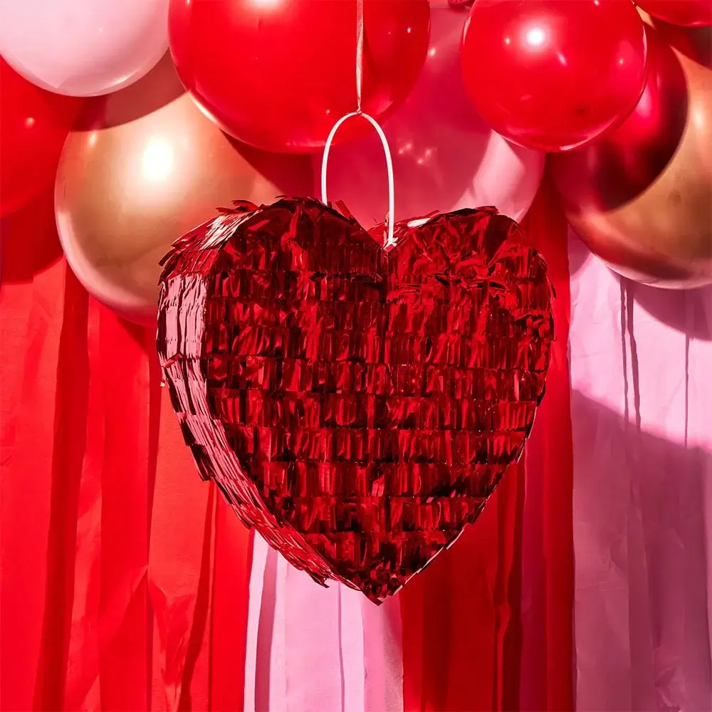Red heart-shaped piñata hanging in front of red, pink, and gold balloons with matching streamers in the background.